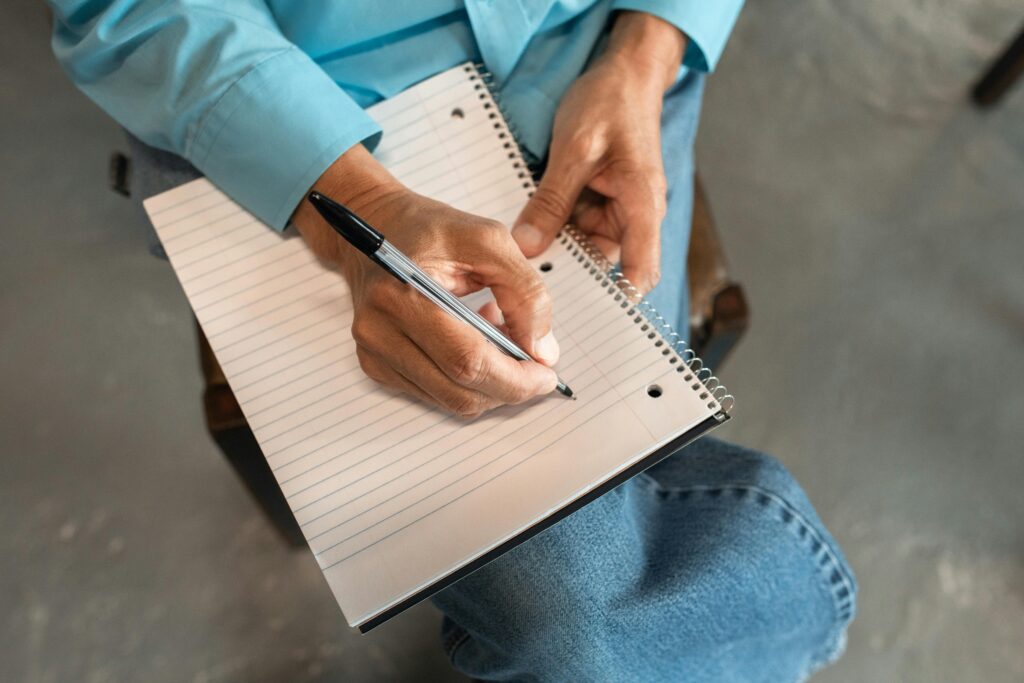 Close-up of hands writing in a spiral notebook with a pen, seated comfortably.